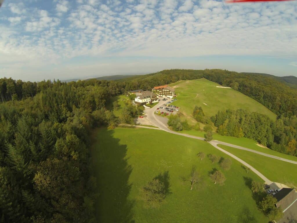 Eine Luftaufnahme einer ländlichen Landschaft zeigt eine malerische Kulisse, die sich perfekt für Hochzeitsfeiern eignet. Ein großes Gebäude, ideal für Hochzeitsplanung, liegt eingebettet zwischen grünen Feldern und dichtem Wald. Eine kurvenreiche Straße führt zu diesem bezaubernden Ort, wobei der teilweise bewölkte Himmel vereinzelte Schatten wirft.