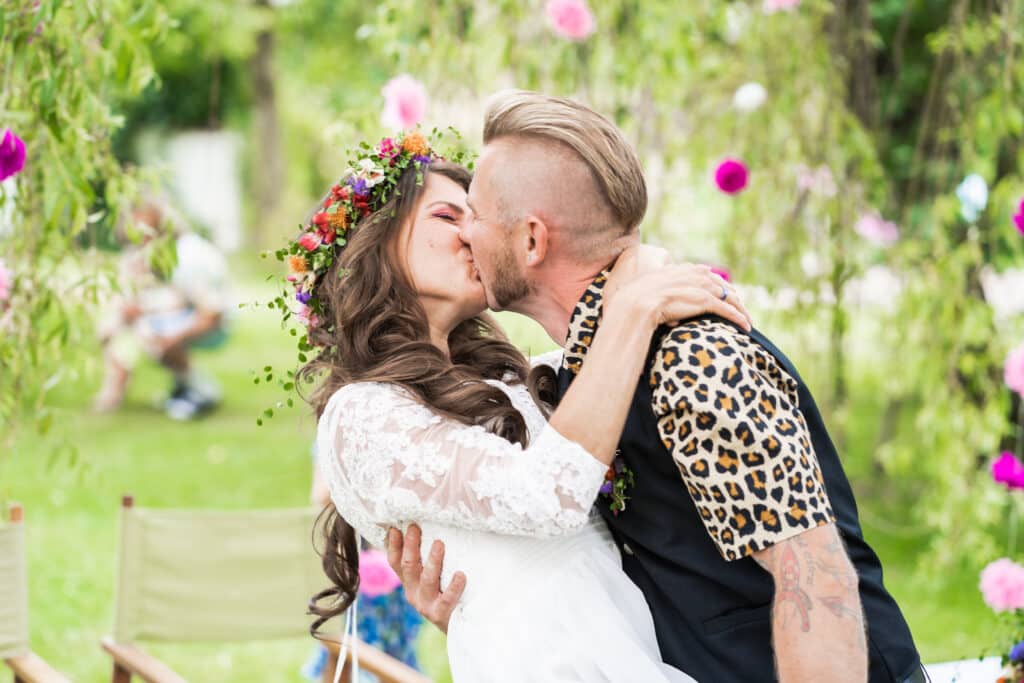 Ein Paar küsst sich freudig im Freien inmitten üppiger Vegetation und bunter Blumen und feiert seine Hochzeit. Die Frau trägt ein weißes Kleid und einen Blumenkranz, was perfekt zum Anlass passt, während der Mann ein Hemd und eine Weste mit Leopardenmuster trägt. Sie umarmen sich eng und genießen diesen Moment der Liebe.