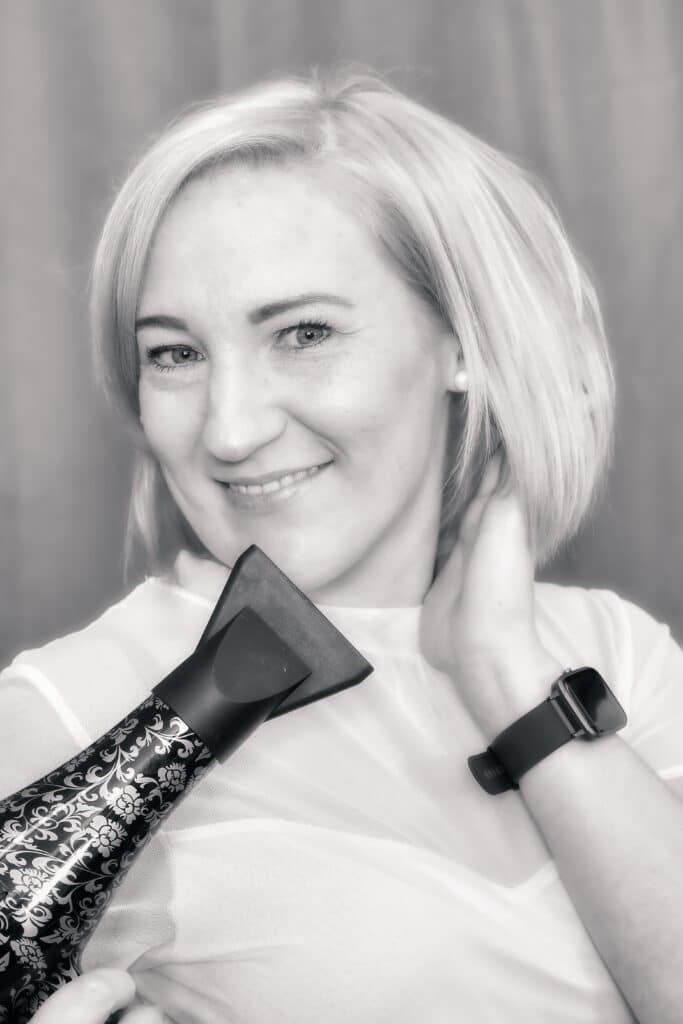Eine Frau mit kurzen blonden Haaren hält einen Haartrockner an ihren Kopf. Sie lächelt und trägt ein helles Oberteil und ein dunkles Armband. Sie bereitet sich wahrscheinlich auf ihre Hochzeit vor. Das Bild ist in Schwarzweiß gehalten und steht vor einem schlichten Hintergrund.