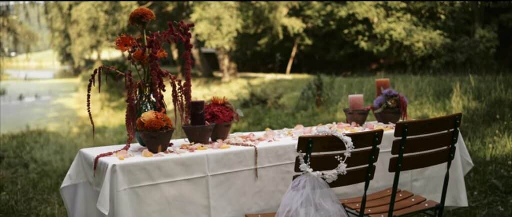 Ein mit einer weißen Tischdecke bedeckter Tisch im Freien mit Blumenarrangements, Kerzen und verstreuten Blütenblättern fängt die Essenz einer verträumten Hochzeit ein. Am Tisch stehen zwei Holzstühle, einer davon mit einem Schleier geschmückt. Der Hintergrund zeigt üppiges Grün und Bäume, perfekt für exquisite Hochzeitsplanung.