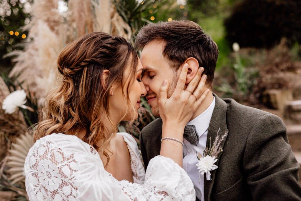 Ein Paar in Hochzeitskleidung teilt einen intimen Moment im Freien. Die Frau im weißen Spitzenkleid hält sanft das Gesicht des Mannes, während sie sich nah aneinanderlehnen, umgeben von Grün und sanftem Licht – eine perfekte Szene für diejenigen, die von ihrem eigenen Hochzeitstag träumen.