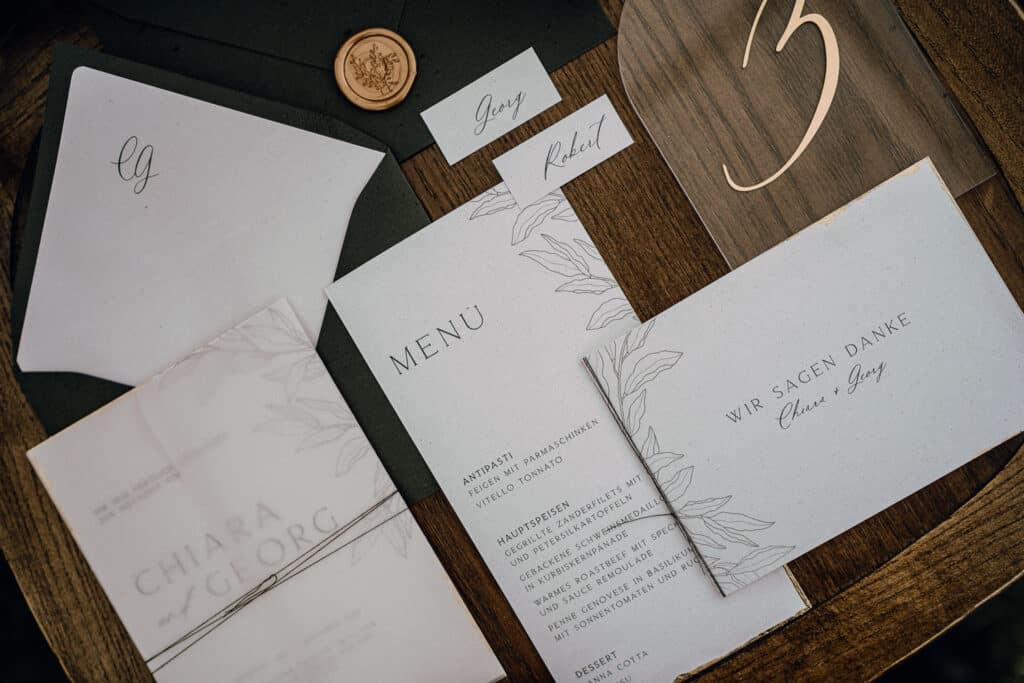 Eine Sammlung eleganter Hochzeitspapeterie, darunter ein Menü, eine Dankeskarte und eine Einladung mit Blumenmustern. Perfekt für die Hochzeitsplanung, jedes Stück wird von einem Wachssiegel und einer personalisierten Karte mit „Greg“ darauf begleitet. Das Set wird wunderschön auf einer Holzoberfläche präsentiert.