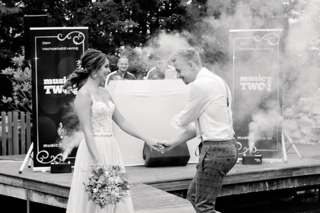 Ein lächelndes Brautpaar tanzt fröhlich im Freien auf einer Holzbühne und feiert seine Hochzeit. Der Bräutigam trägt Hemd und Hosenträger, während die Braut ihren Brautstrauß hält. Im Hintergrund erzeugen Lautsprecher und ein DJ einen bezaubernden Nebel, der ihrer perfekten Hochzeitsplanung Magie verleiht.