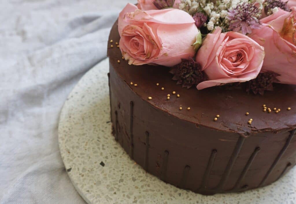 Ein Schokoladenkuchen mit geschmeidiger, reichhaltiger Glasur ist oben mit rosa Rosen und kleinen Blumen geschmückt. Perfekt für eine Hochzeit, mit zarten Goldperlen und elegant auf einem gesprenkelten weißen Teller – ein ideales Herzstück für jede Hochzeitsfeier.