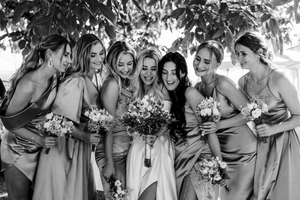 Ein Schwarzweißfoto einer Gruppe von Frauen in eleganten Kleidern, die freudig zusammengekauert sind. Sie halten kleine Blumensträuße in den Händen und stehen unter einem Baum, über dem die Blätter ein natürliches Blätterdach bilden – ein perfekter Moment, festgehalten während einer Hochzeitsfeier.