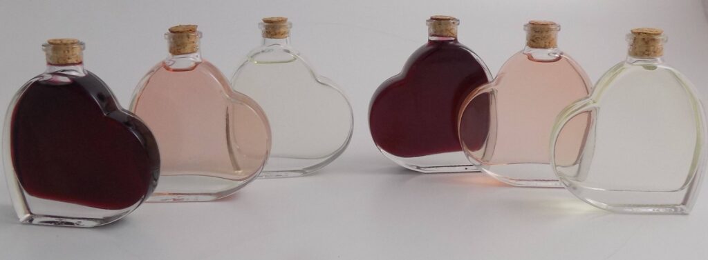 Sechs herzförmige Glasflaschen mit Korkverschluss, perfekt, um jeder Hochzeit eine skurrile Note zu verleihen. Jede Flasche ist mit Flüssigkeiten in verschiedenen Farben gefüllt, darunter Dunkelrot, Hellrosa und Klar, elegant in einer Reihe auf einer weißen Oberfläche angeordnet. Ideal für Ihre Traumhochzeitsplanung!