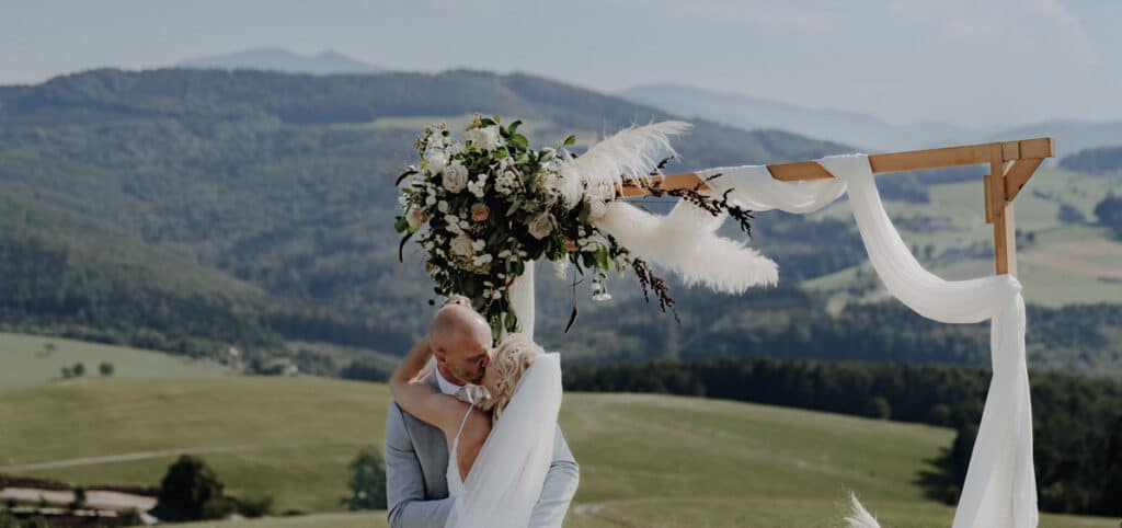 Ein Paar umarmt sich unter einem hölzernen Hochzeitsbogen, der mit Blumen und weißem Stoff geschmückt ist, vor der Kulisse sanfter grüner Hügel und Berge in der Ferne unter einem blauen Himmel – ein malerischer Moment ihrer Hochzeit.