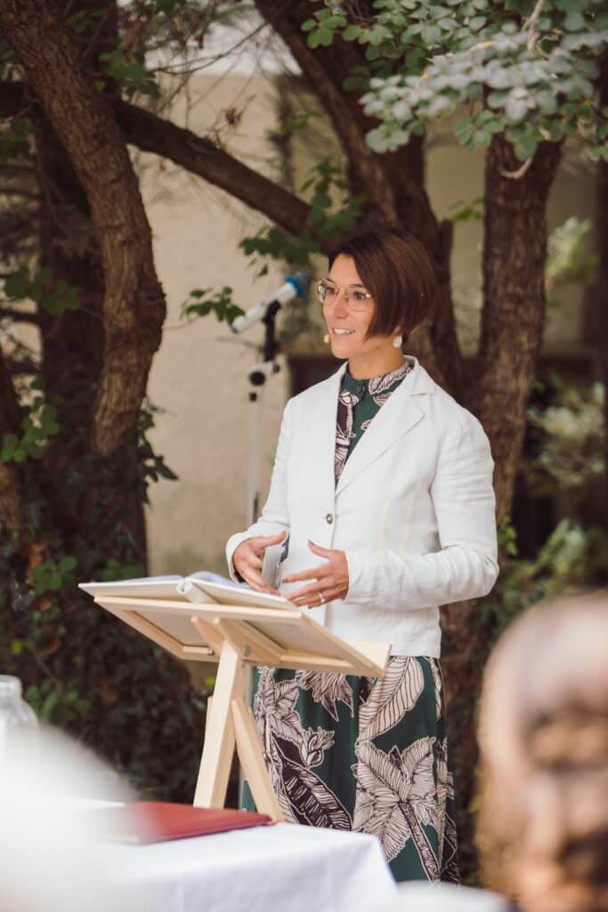 An einem malerischen Veranstaltungsort im Freien, umgeben von üppigem Grün, hält eine Person in einem weißen Blazer und einem Kleid mit Blumenmuster eine eloquente Rede am Podium. Ihre Brillengläser glitzern im Sonnenlicht und verleihen dieser bezaubernden Hochzeitsfeier einen Hauch von Eleganz.
