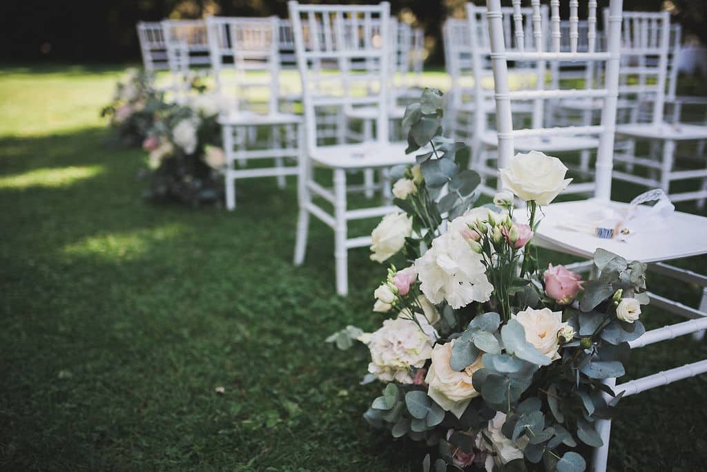 Weiße Stühle, in Reihen auf einer Rasenfläche angeordnet, geschmückt mit Blumenarrangements aus weißen und rosa Rosen mit Eukalyptusblättern, was den Eindruck einer Hochzeit oder Zeremonie im Freien erweckt.