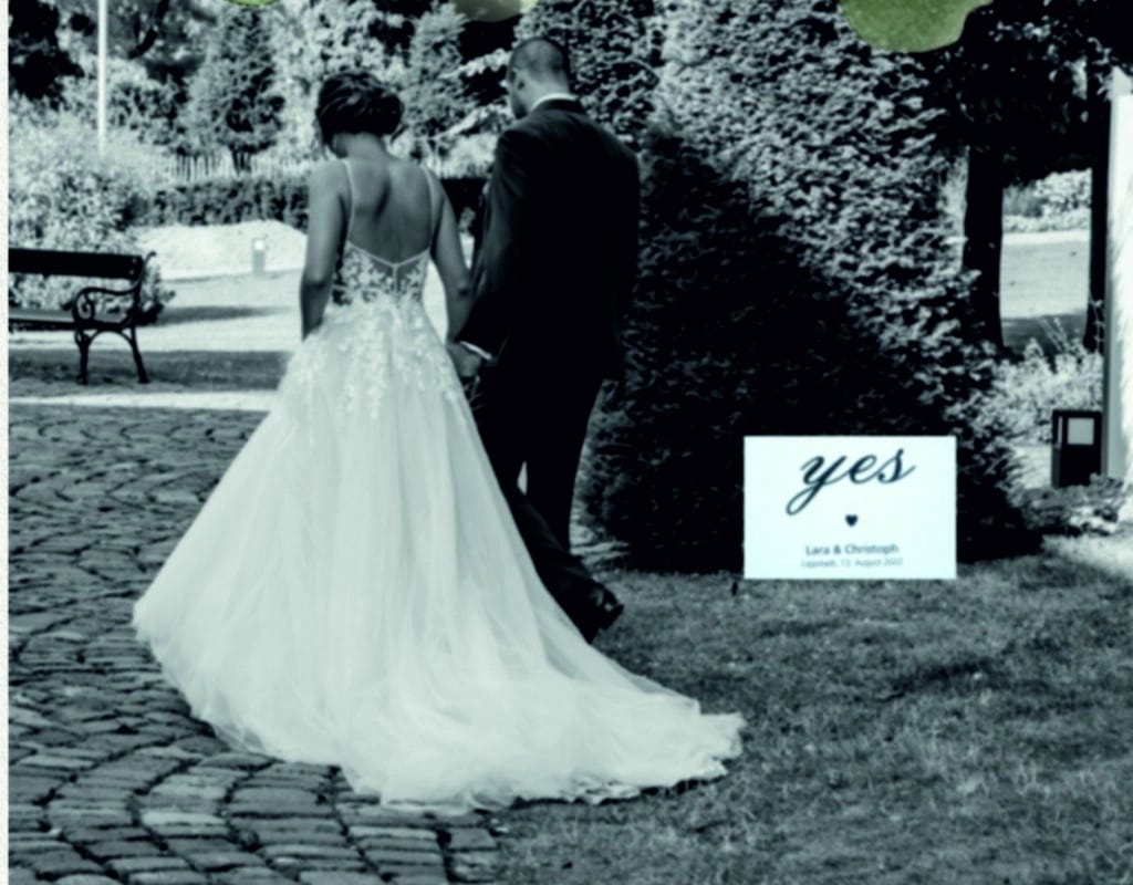 Ein Schwarzweißfoto zeigt ein Paar, das Hand in Hand einen Kopfsteinpflasterweg entlang geht. Die Braut in ihrem wallenden Kleid und der Bräutigam in seinem Anzug verkörpern die Essenz von „heiraten“. Auf einem Schild daneben steht „Ja“, darunter steht elegant „Lara & Christoph“.