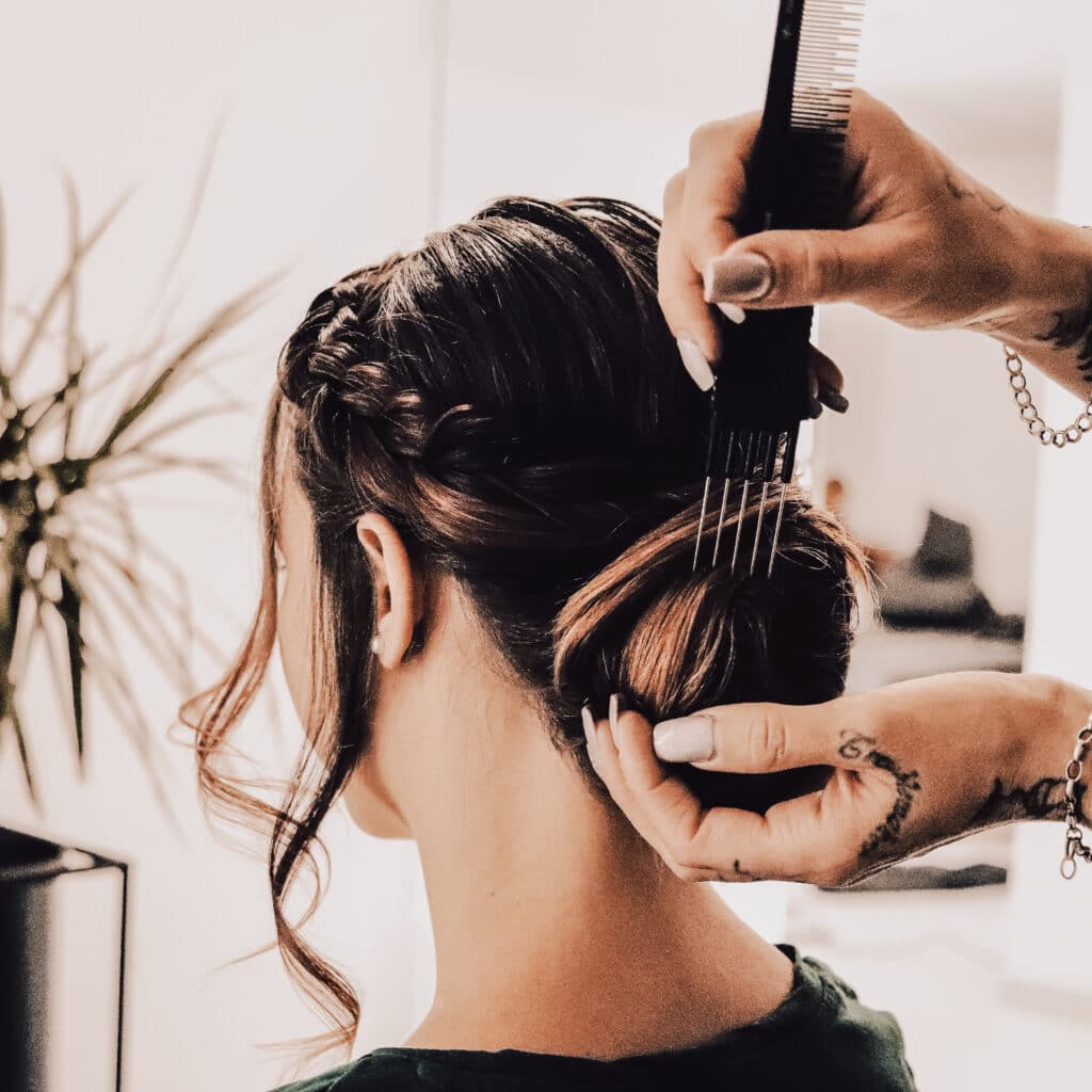Eine Person mit braunem Haar sitzt da, während eine andere ihr Haar mithilfe eines Kamms fachmännisch zu einer eleganten Hochsteckfrisur für eine Hochzeit frisiert. Der gestylte Look umfasst Zöpfe und weiche Locken, perfekt für einen solchen Anlass. Eine grüne Pflanze trägt im unscharfen Hintergrund zur ruhigen Atmosphäre bei.