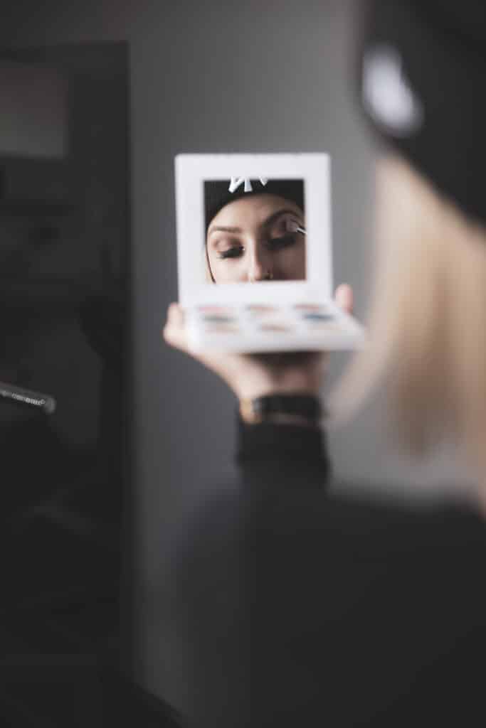 Eine Person mit dunklem Hut trägt Make-up und blickt dabei in einen kleinen Handspiegel, während sie sich sorgfältig auf ihre Hochzeit vorbereitet. Der Fokus liegt auf ihrem Auge, eingefangen in dem sanft beleuchteten Raum, während sie sich darauf vorbereitet, Liebe und Heirat zu feiern.