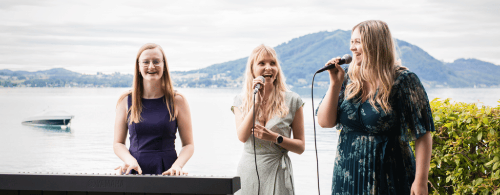 Drei Frauen treten bei einer wunderschönen Hochzeit am Seeufer auf; eine spielt Keyboard, während zwei in Mikrofone singen. Sie stehen vor einer malerischen Kulisse mit einem ruhigen See und fernen Hügeln unter einem bewölkten Himmel und schaffen eine bezaubernde Atmosphäre für die Feier.