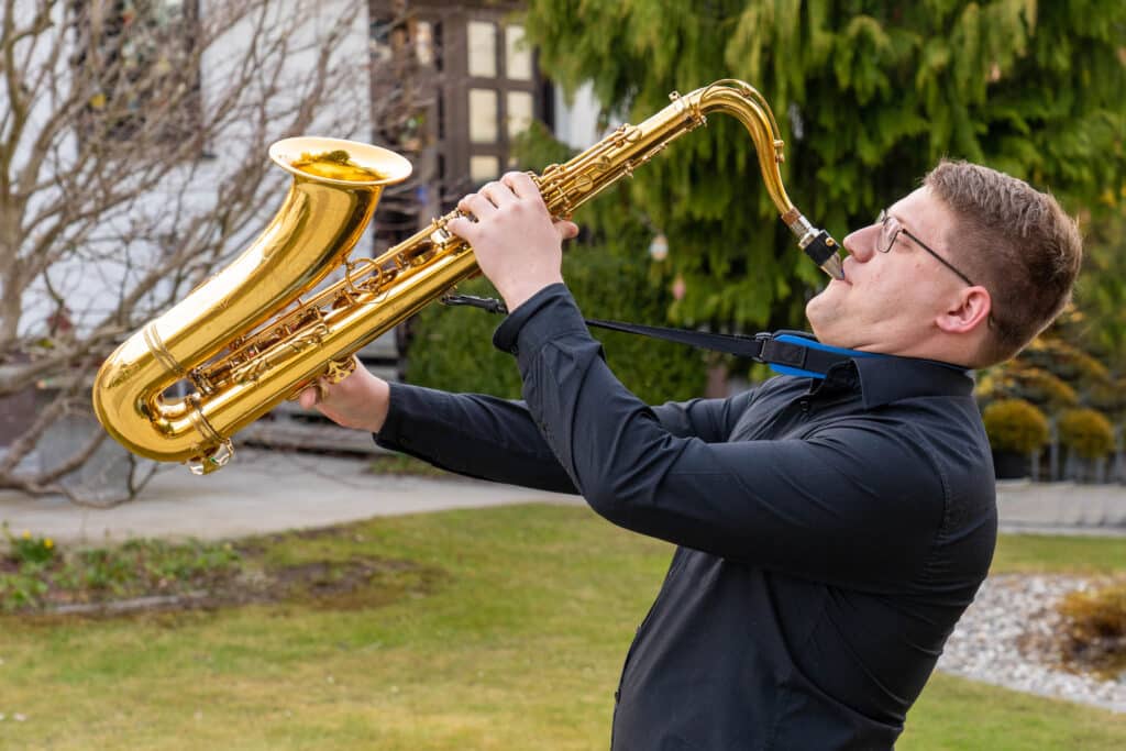 Inmitten der ruhigen Außenkulisse spielt ein Mann in schwarzem Hemd und Brille leidenschaftlich ein goldenes Baritonsaxophon, sein Blick ist nach oben gerichtet, als würde er den Bäumen und dem Gebäude ein Ständchen singen. Die Szene ruft eine Atmosphäre der Harmonie hervor, die an eine wunderschön orchestrierte Hochzeit erinnert.