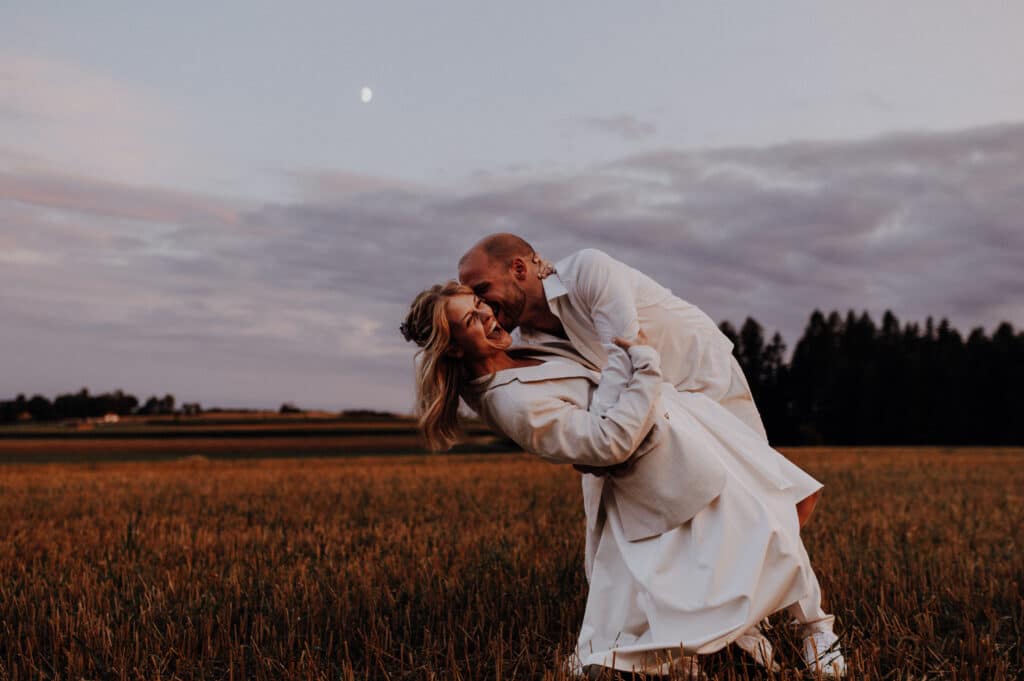 Ein Mann taucht eine Frau bei Sonnenuntergang spielerisch in ein Feld und fängt damit die freudige Stimmung ihrer Hochzeit ein. Beide sind in Weiß gekleidet, über ihnen ist der Mond am bewölkten Himmel zu sehen. Die Szene ruft Romantik und Freude vor einem Hintergrund aus offenen Feldern und Bäumen hervor, perfekt für Heiratsträume.