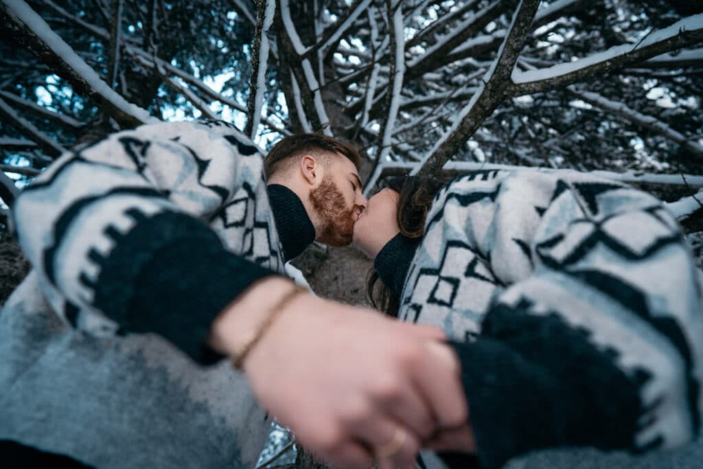 Ein Paar in passenden gemusterten Pullovern küsst sich unter einer schneebedeckten Kiefer, aufgenommen aus niedriger Perspektive. Der Schnee liegt sanft auf den Zweigen darüber und verleiht eine romantische Note, die perfekt zu ihren Hochzeitsplanungsträumen passt.