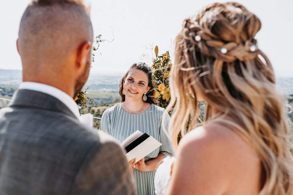 Ein Paar steht mit dem Rücken zur Kamera und blickt auf eine Frau, die ihre Hochzeit im Freien durchführt. Sie sind von Sonnenlicht und Grün umgeben, mit einer malerischen Landschaft im Hintergrund. Die Trauzeugin lächelt und hält ein Buch in der Hand, was diesem wunderschönen Moment der Hochzeit Wärme verleiht.