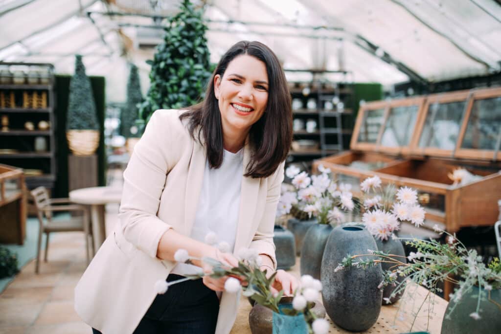Eine Frau lächelt, während sie Blumen in einem Gewächshaus arrangiert, und träumt vielleicht von ihrer eigenen Hochzeit. Sie trägt einen hellen Blazer über einem weißen Oberteil. Im Hintergrund sind verschiedene Pflanzen, Blumen und Holzmöbel zu sehen, perfekt für jede Hochzeitsdeko-Inspiration.