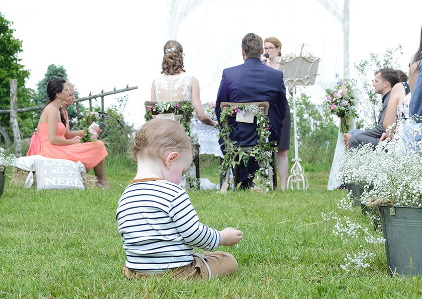 Ein Kind sitzt bei einer Hochzeitszeremonie im Freien auf dem Rasen. Im Hintergrund sitzt ein Paar gegenüber dem Geistlichen. Die Gäste sitzen um sie herum und Blumenschmuck schmückt die Szene. Die Atmosphäre ist heiter und natürlich.