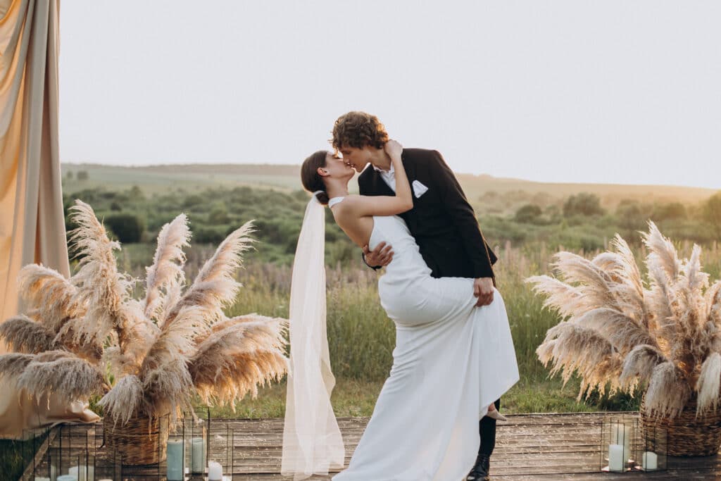 Ein Paar im Hochzeitskleid küsst sich romantisch in einer malerischen Umgebung im Freien bei Sonnenuntergang. Sie sind von dekorativem Pampasgras und einem goldenen Vorhang im Hintergrund umgeben und verkörpern die Magie einer Hochzeit mit der wunderschönen Landschaft, die einen unvergesslichen Moment malt.