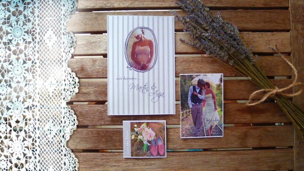 Eine Hochzeitseinladung auf einem Holztisch, bestehend aus einer Karte mit einem Bild von Braut und Bräutigam, einem Strauß rosa und weißer Blumen und einem Foto des Paares. Ein Bündel Lavendel und Spitzenstoff ergänzen die elegante Präsentation, perfekt für alle, die gerade mitten in der Hochzeitsplanung stecken.