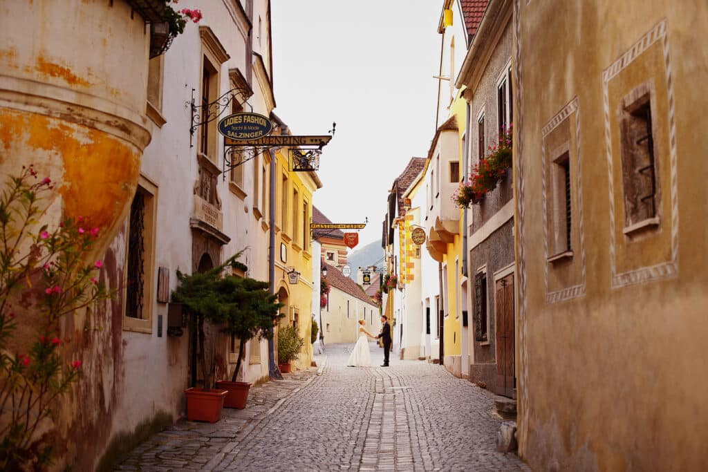 Eine schmale, gepflasterte Straße, gesäumt von farbenfrohen, rustikalen Gebäuden, die mit hängenden Schildern und Topfpflanzen geschmückt sind. Ein Paar in Abendgarderobe, das wahrscheinlich seine Hochzeit plant, geht Hand in Hand die charmante Gasse entlang.