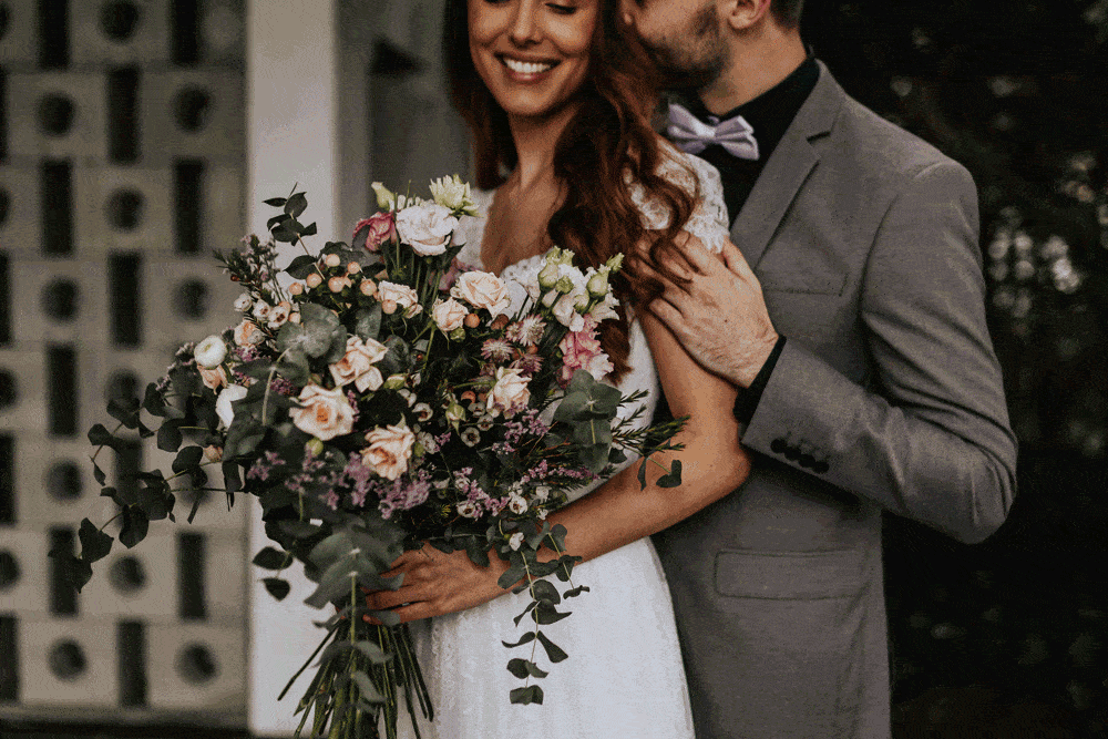 Eine Braut mit einem großen Blumenstrauß steht neben einem Bräutigam in einem grauen Anzug mit Fliege und verkörpert damit die Essenz einer Hochzeit. Der Bräutigam hat seinen Arm um sie gelegt und flüstert ihr etwas ins Ohr. Beide lächeln herzlich, eingerahmt von einem sanft verschwommenen Hintergrund, der ihren intimen Heiratsmoment einfängt.