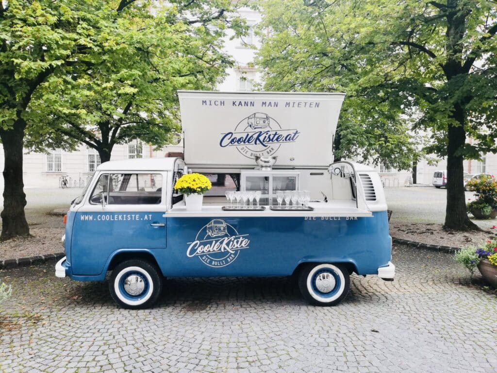 Ein blauer Oldtimer-Van dient als mobile Bar und steht auf einem Kopfsteinpflasterweg unter grünen Bäumen. Perfekt für alle, die ihrer Hochzeit eine charmante Note verleihen möchten. Der „Coole Kiste“-Van präsentiert Glaswaren und eine gelbe Blume in der offenen Heckklappe und schafft so eine malerische und einladende Atmosphäre.