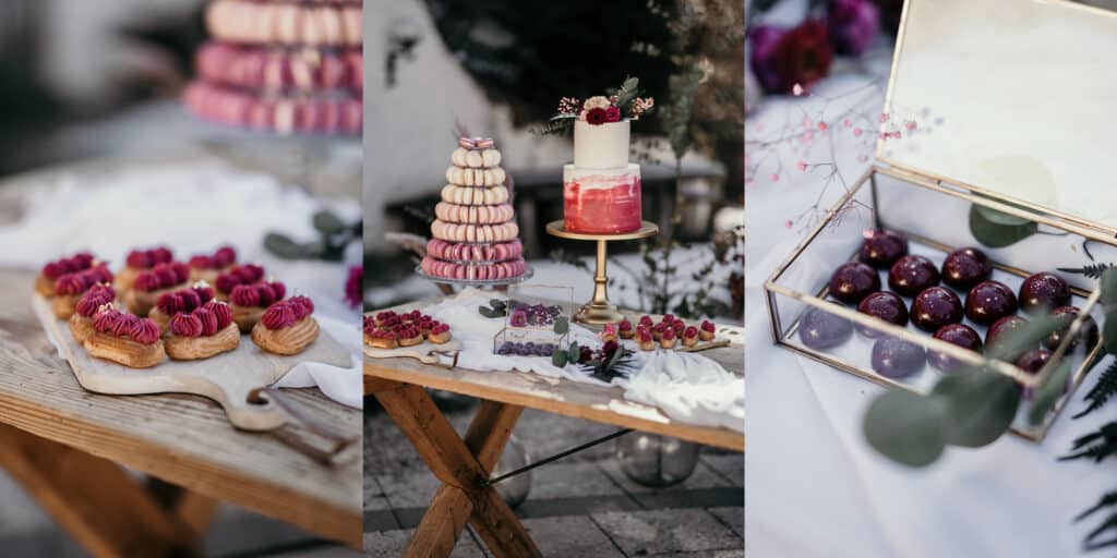 Ein Desserttisch, der perfekt zu einer Hochzeit passt: Gebäck mit rosa Creme, ein mehrstöckiger Macaron-Turm und ein rot-weißer Kuchen, der mit Blumen geschmückt ist. Quadratische Süßigkeiten und mit Schokolade überzogene Köstlichkeiten füllen eine Glasbox. Die Außenkulisse bietet ein charmantes rustikales Dekor, ideal für Hochzeitsfeiern.