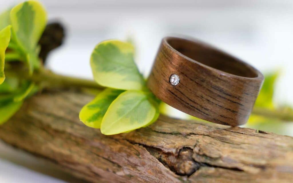 Ein Holzring mit einem kleinen Diamanten ist kunstvoll auf Treibholz platziert, umgeben von üppigen grünen Blättern. Perfekt für diejenigen, die gerade mitten in der Hochzeitsplanung stecken. Seine natürliche Textur und die erdige Fassung betonen sein einzigartiges Design und machen ihn zu einem idealen Symbol für Liebe und Verbundenheit.