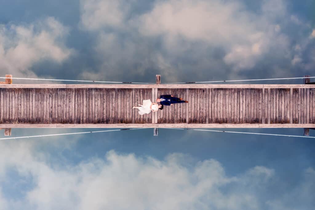 Eine Luftaufnahme von Braut und Bräutigam, die über eine Holzbrücke über ruhiges Wasser laufen, in dem sich Himmel und Wolken spiegeln. Die Braut trägt ein weißes Kleid, der Bräutigam einen dunklen Anzug. Die ruhige Umgebung schafft eine verträumte Atmosphäre, perfekt für eine Hochzeit, die für immer in Erinnerung bleiben wird.