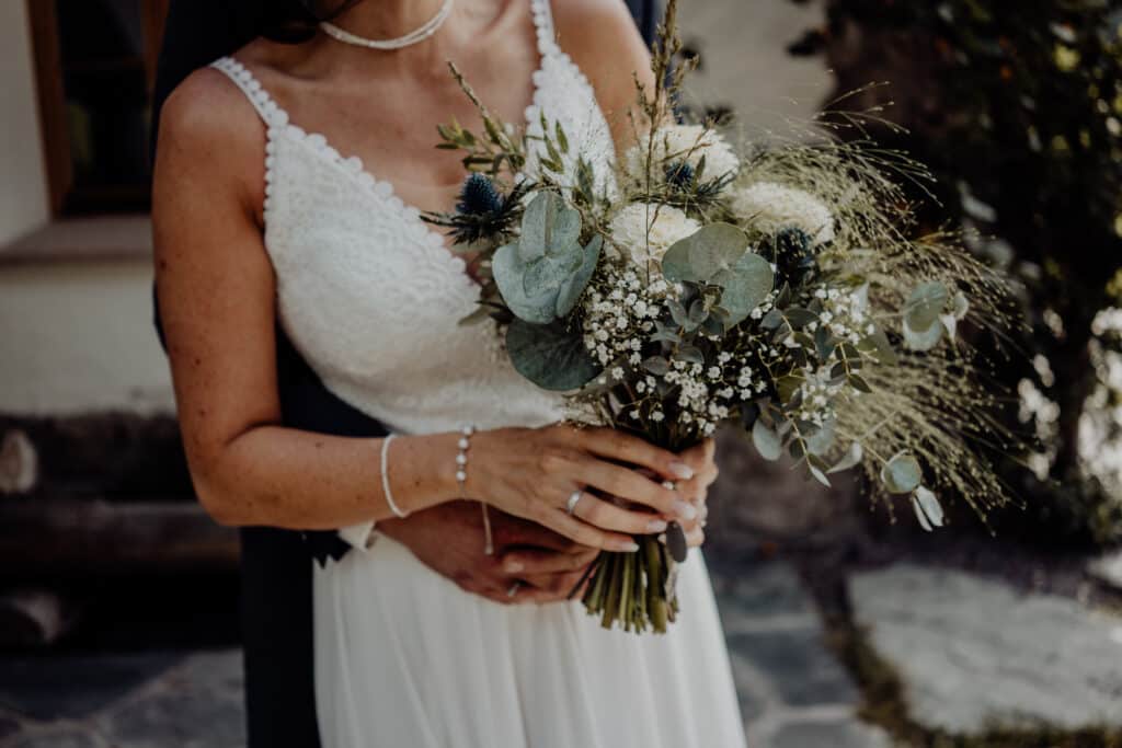 In einer rustikalen Umgebung im Freien hält eine Braut in einem weißen Spitzenkleid einen Strauß aus Grünpflanzen und weißen Blumen. Von hinten sanft umarmt, fängt der Moment die Essenz ihrer Hochzeitsreise ein.