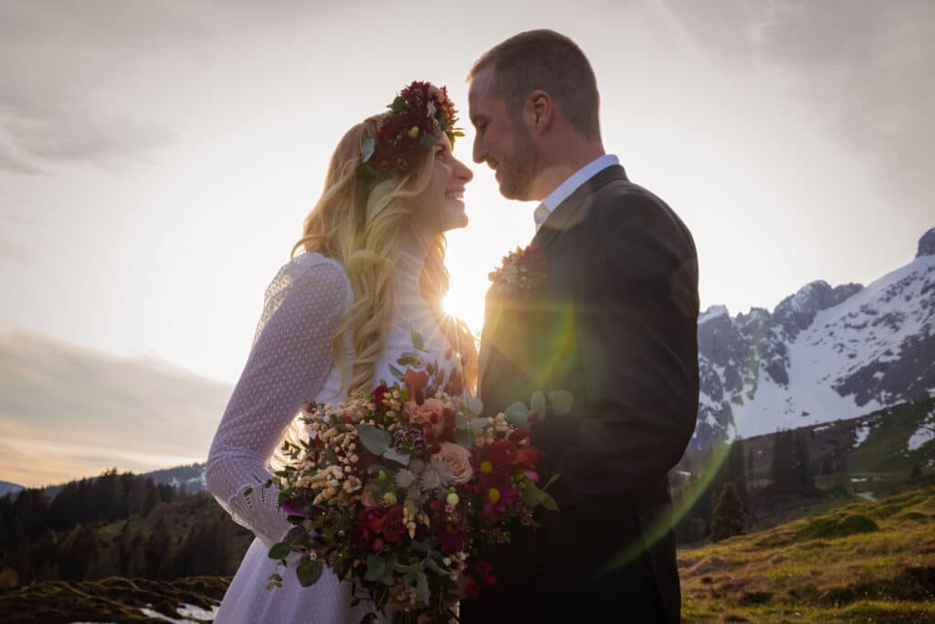 Ein Paar umarmt sich bei Sonnenuntergang vor einer Berglandschaft und verkörpert die Romantik einer perfekten Hochzeit. Die Frau in einem weißen Kleid und einem Blumenkopfschmuck hält einen Blumenstrauß. Sonnenlicht fällt mit einem warmen Schein zwischen sie hindurch, während schneebedeckte Gipfel ihren Moment einrahmen.