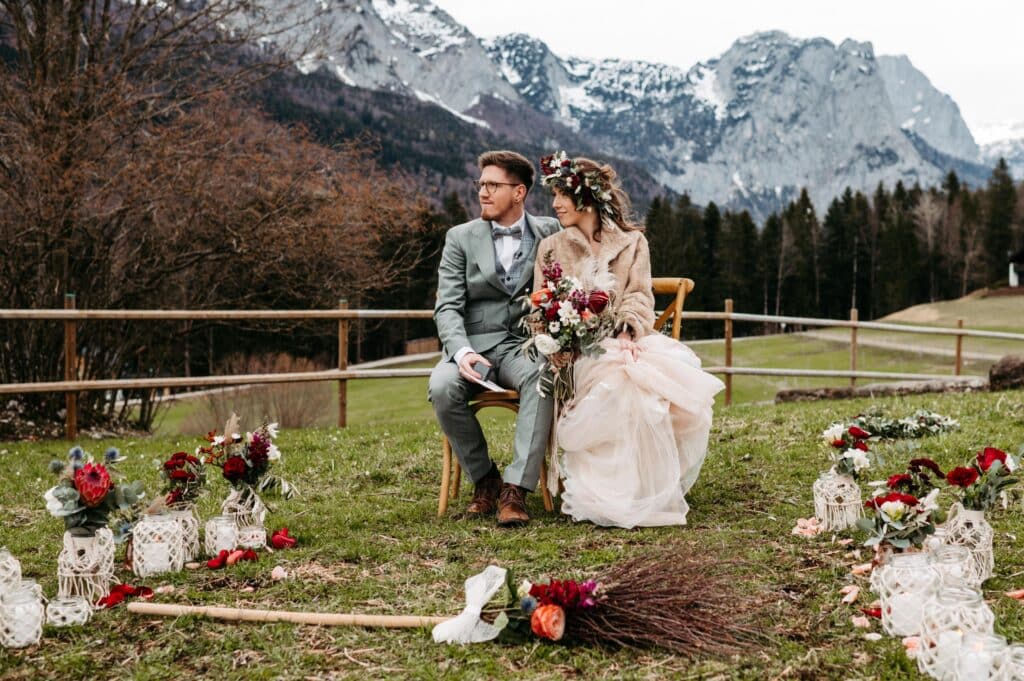 Ein Paar sitzt in Hochzeitskleidung auf einem Stuhl in einer Berglandschaft. Die Braut hält einen Blumenstrauß und Blumendekorationen mit Laternen umgeben sie und zeigen perfekte Hochzeitsplanung. Im Hintergrund sind schneebedeckte Gipfel und Tannenbäume zu sehen, die den Zauber des Heiratens inmitten der Natur einfangen.
