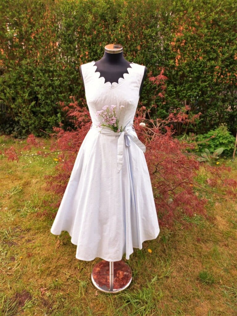 Eine Schaufensterpuppe präsentiert ein hellblaues, ärmelloses Kleid mit gewelltem Ausschnitt und Gürtel in der Taille, geschmückt mit kleinen Lavendelblüten – perfekt für eine romantische Hochzeit. Der Hintergrund aus grünem und rotem Laub unterstreicht die verträumte Gartenkulisse.