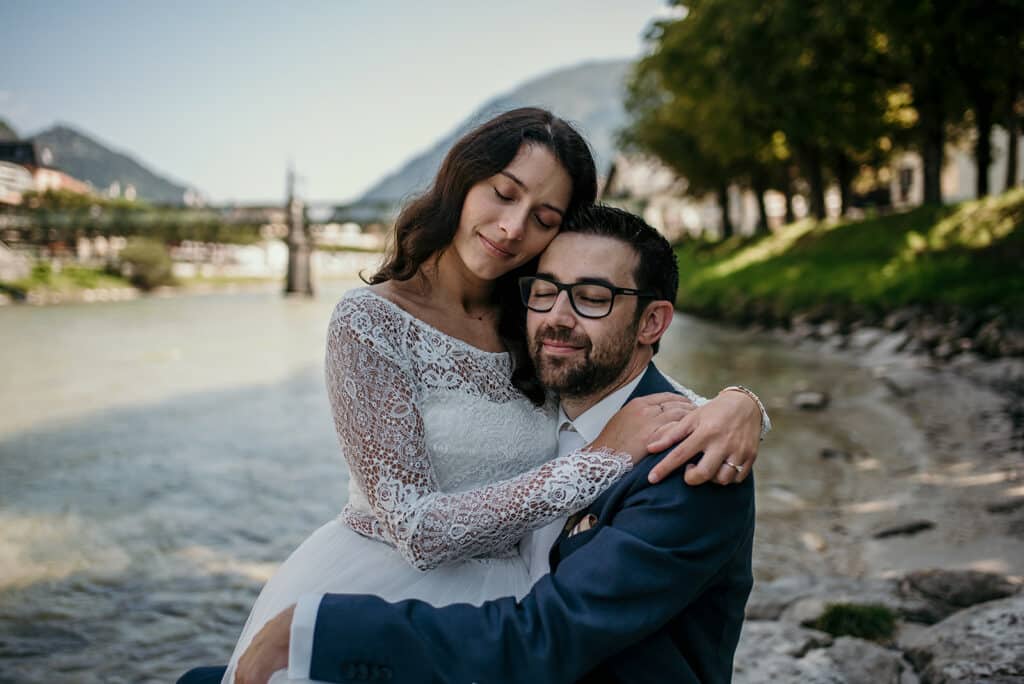 Braut und Bräutigam umarmen sich an einem ruhigen Fluss, umgeben von Bäumen und Bergen, und verkörpern so ihre Traumhochzeit. Sie trägt ein Spitzenbrautkleid und er einen dunklen Anzug mit Brille. Beide lächeln mit geschlossenen Augen und genießen die Ruhe dieses perfekt geplanten gemeinsamen Moments.
