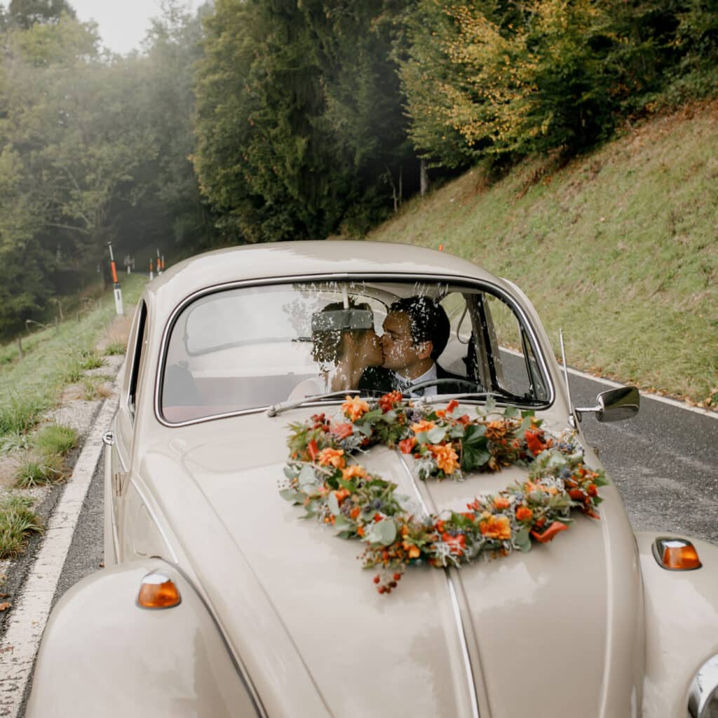 Ein Paar küsst sich in einem cremefarbenen Oldtimer mit einem farbenfrohen Blumenkranz auf der Motorhaube und feiert seine Hochzeit. Sie fahren auf einer malerischen Straße, umgeben von üppigem Grün und Bäumen, was die Essenz ihrer romantischen Reise einfängt.