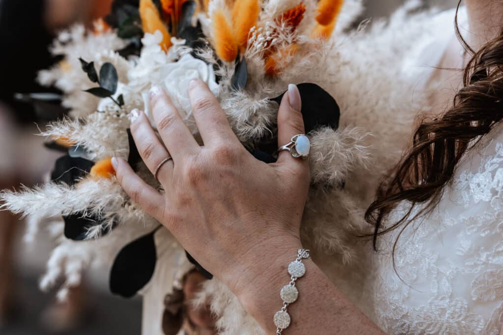 Die Hand einer Frau, geschmückt mit einem silbernen Armband und einem Ring mit einem blauen Stein, hält elegant einen Strauß getrockneter Blumen und weißer Rosen. Perfekt für jede Hochzeit, fängt die Szene die romantische Essenz des Heiratens vor einem wunderschön verschwommenen Hintergrund ein.