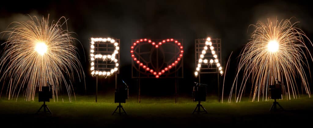 Ein helles Feuerwerk bildet ein Herzsymbol zwischen den Buchstaben „B“ und „A“ mit funkelnden Explosionen auf beiden Seiten vor einem dunklen Hintergrund und schafft so eine unvergessliche Szene für Ihre Hochzeitsplanung.