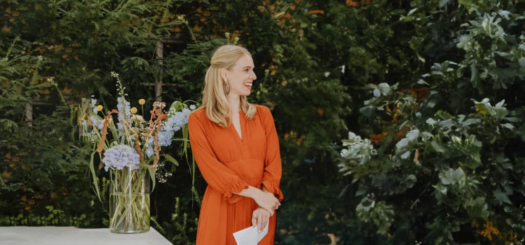 Eine Frau in einem orangefarbenen Kleid steht draußen neben einem großen Strauß bunter Blumen und strahlt die Freude ihrer Hochzeit aus. Sie lächelt, umgeben von üppigem grünem Laub, und fängt die glückselige Essenz sorgfältiger Hochzeitsplanung perfekt ein.