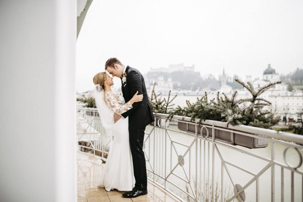 Braut und Bräutigam küssen sich auf einem Balkon mit Blick auf eine neblige Stadtlandschaft und feiern ihre Hochzeit. Die Braut trägt ein weißes Kleid mit Spitzenärmeln, während der Bräutigam einen schwarzen Anzug trägt. Grün schmückt die intime Szene, in der elegante Hochzeitsplanung ihren besonderen Tag akzentuiert.