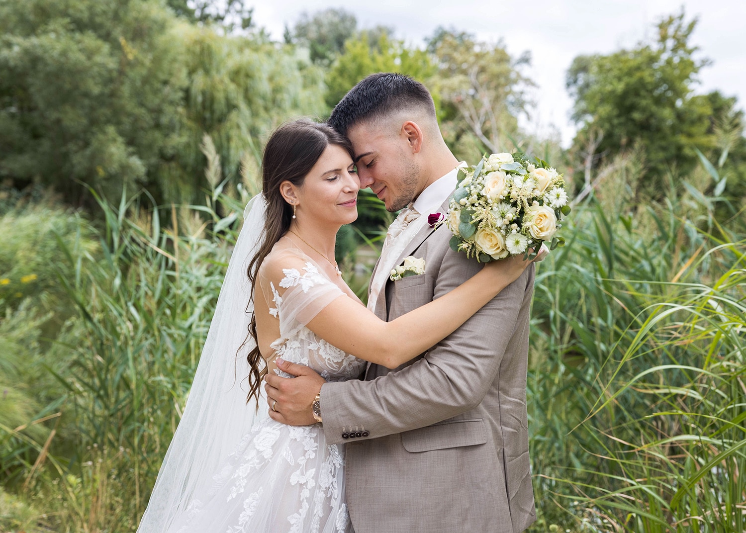 Braut und Bräutigam umarmen sich im Freien und feiern ihre Hochzeit. Die Braut in einem weißen Spitzenkleid hält einen Strauß cremefarbener und weißer Blumen. Der Bräutigam trägt einen beigen Anzug. Sie stehen inmitten üppigen Grüns und vermitteln eine heitere und romantische Atmosphäre.