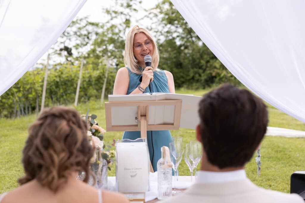Eine Frau mit blonden Haaren, Brille und blauem Kleid steht an einem Rednerpult und spricht unter einem weißen Baldachin in ein Mikrofon. Es ist Teil einer eleganten Hochzeitsszene. Im Vordergrund sitzen zwei Personen an einem Tisch, auf dem Champagnergläser und eine Speisekarte liegen. Im Hintergrund sind Grünpflanzen zu sehen.