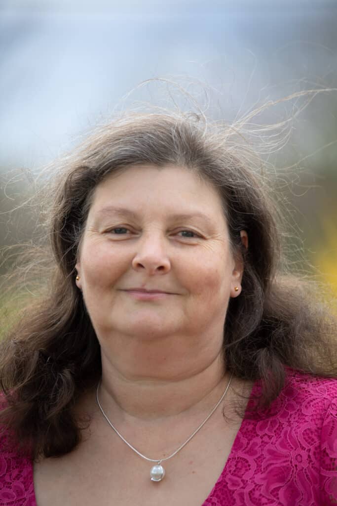 Eine Frau mit langen braunen Haaren und einem sanften Lächeln trägt ein rosa Spitzentop und eine Halskette mit Perlenanhänger und verströmt damit einen Hauch von Romantik, der perfekt für die Hochzeitsplanung ist. Der Hintergrund ist sanft unscharf.