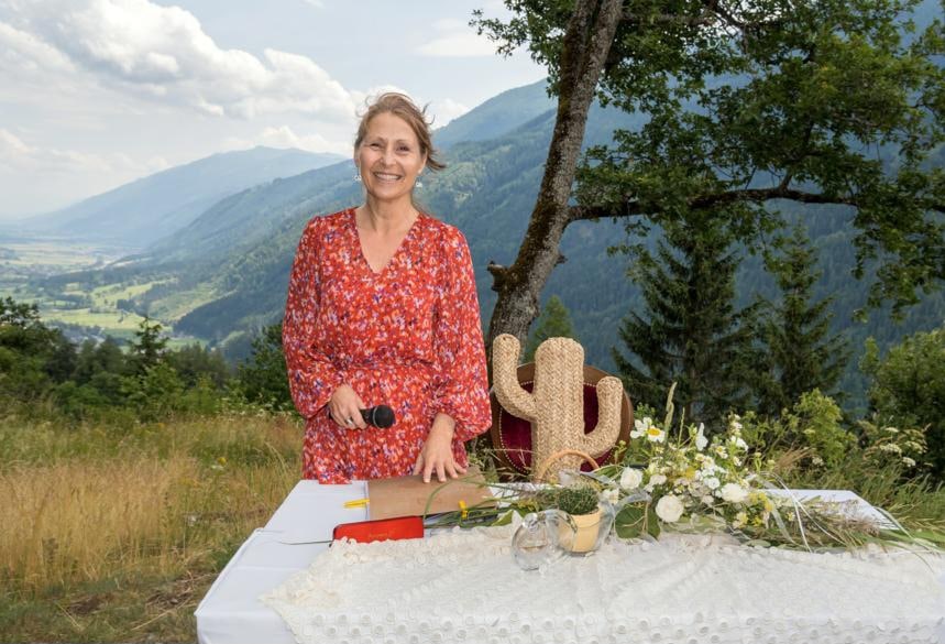 Eine Frau in einem roten Blumenkleid steht lächelnd hinter einem Tisch mit einer weißen Spitzendecke, perfekt für die Hochzeitsplanung. Auf dem Tisch liegt ein Strauß weißer Blumen und ein dekorativer Kaktus, während Berge und Grün die malerische Kulisse bilden.