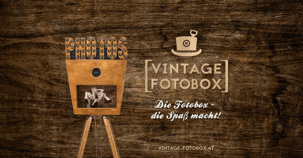 Eine charmante Vintage-Fotobox aus Holz, ideal für die Hochzeitsplanung, verfügt über ein Kameraobjektiv und einen Monitor, auf dem zwei Personen abgebildet sind. Daneben ist der Text „Vintage Fotobox“ mit einem Zylinder-Logo verziert. Die satte braune Holzstruktur des Hintergrunds trägt zur bezaubernden Hochzeitsatmosphäre bei.