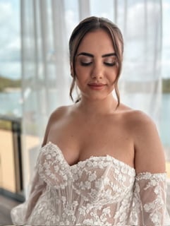 Eine Frau in einem weißen Spitzenbrautkleid, perfekt für jede Hochzeitsplanung, steht vor transparenten Vorhängen. Ihre Augen sind geschlossen und ihr Haar ist hochgesteckt, wobei Strähnen ihr Gesicht umrahmen. Natürliches Licht erhellt die ruhige Szene wunderschön.