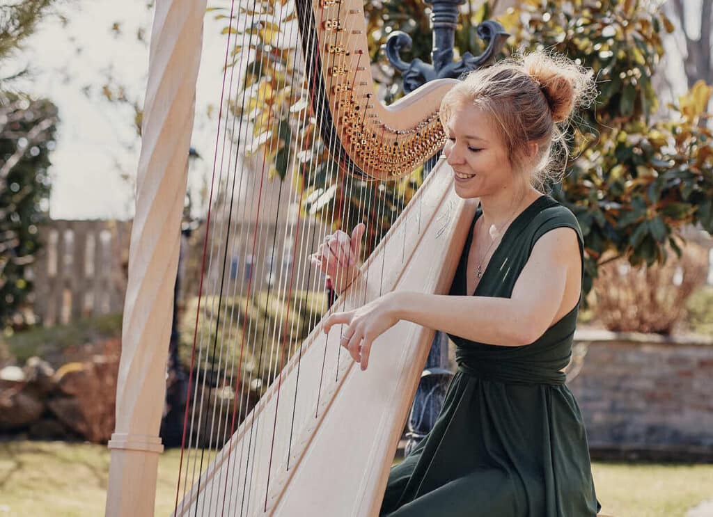 Eine Frau in einem grünen Kleid spielt im Freien, umgeben von sonnigem Grün, Harfe. Ihr Lächeln trägt zur heiteren und friedlichen Atmosphäre bei und schafft eine zauberhafte Kulisse für alle, die von einer Märchenhochzeit träumen.