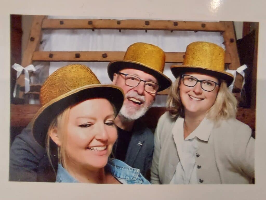 Drei Personen lächeln in die Kamera und tragen goldene Glitzerhüte. Vielleicht feiern sie die erfolgreiche Hochzeitsplanung. In einem Raum mit Holzdekor trägt die Person links eine Jeansjacke, die Person in der Mitte hat einen Bart und die Person rechts trägt eine Brille und eine helle Jacke.