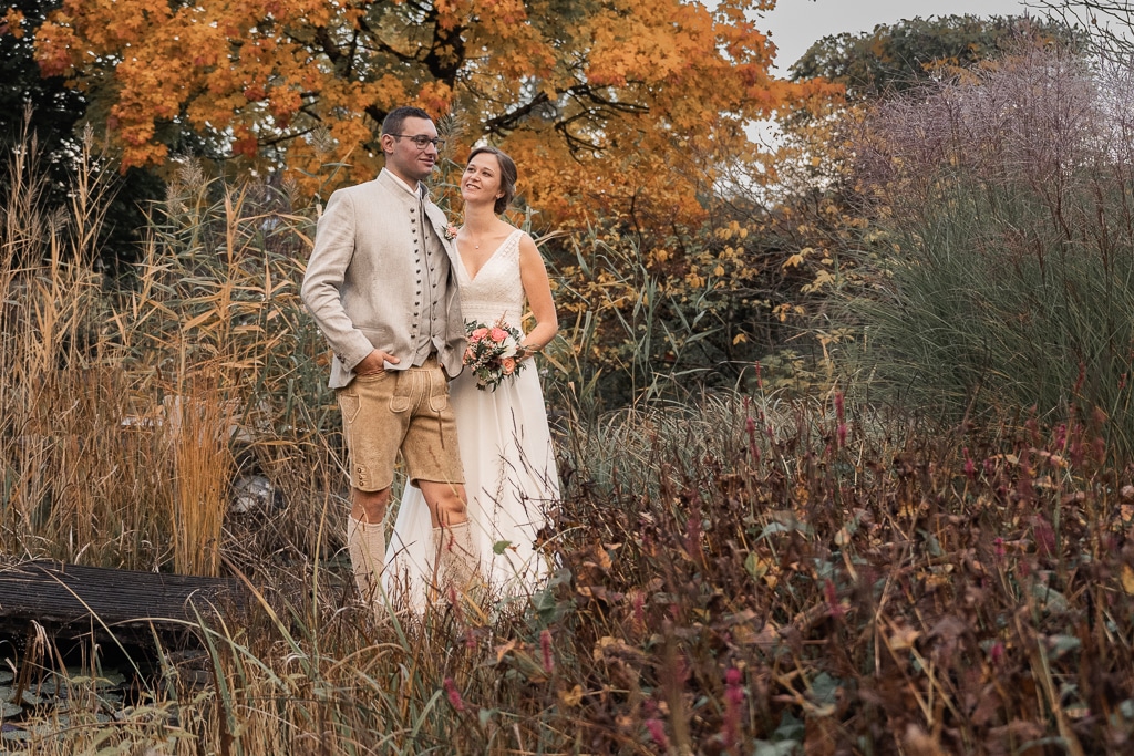 In einem Herbstgarten steht ein Paar inmitten von leuchtendem Laub. Der Mann trägt traditionelle Kleidung mit Jacke und Shorts, während die Frau in ihrem weißen Kleid einen Blumenstrauß hält. Sie lächeln, während sie in die Ferne blicken, ihre Gedanken sind erfüllt von Hochzeitsplanung und Träumen vom Heiraten.